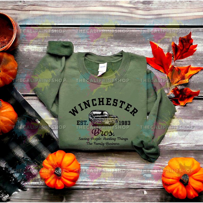 K&ouml;nnte beinhalten: Ein gr&uuml;nes Sweatshirt mit einem schwarzen Grafikdruck eines Autos und dem Text "Winchester Est. 1983 Bros. Saving People Hunting Things The Family Business"