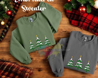 Può includere: Due maglioni di Natale ricamati con un disegno di tre alberi di Natale con luci e stelle. I maglioni sono verde e grigio e sono piegati su una superficie di legno.