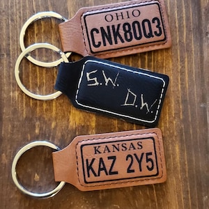 Puede incluir: Tres llaveros de cuero con anillas metálicas. El llavero superior es marrón con el texto "OHIO CNK80Q3". El llavero del medio es negro con el texto "S.W. D.W.". El llavero inferior es marrón con el texto "KANSAS KAZ 2Y5".