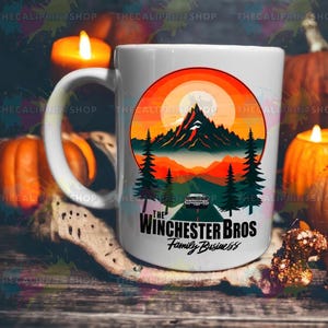 Könnte beinhalten: Weiße Keramik-Tasse mit einem Berglandschaftsdesign, das einen orangefarbenen Sonnenuntergang und Kiefern zeigt. Der Text "The Winchester Bros Family Business" ist unter dem Bild aufgedruckt. Die Tasse hat einen großen Henkel.