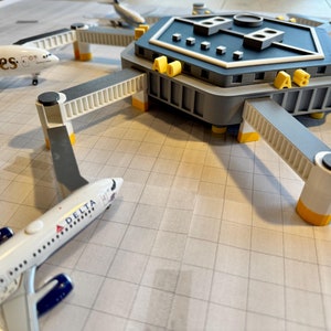 May include: A model airport terminal with six gates, each with a gray and white bridge leading to a plane. The terminal is gray with a white trim and has the letters "A B" on the side. Two planes are visible, one with the word "Emirates" on the side and the other with the word "Delta" on the side.