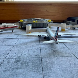 May include: A model airport terminal with three airplanes. A white airplane with red and blue accents is parked at a gate. A white airplane with red accents is parked on the tarmac. A white airplane with blue accents is parked in a hangar.