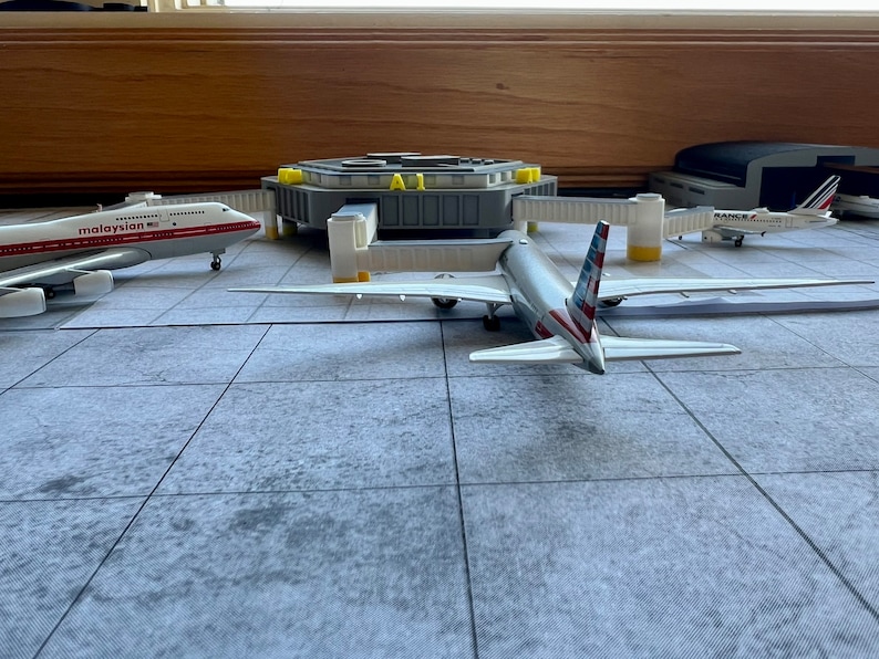 May include: A model airport terminal with three airplanes. A white airplane with red and blue accents is parked at a gate. A white airplane with red accents is parked on the tarmac. A white airplane with blue accents is parked in a hangar.