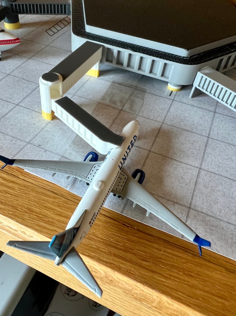 May include: A white and blue airplane model with the word "UNITED" on the side, sitting on a gray and white tiled surface. The airplane is connected to a white and gray gate.