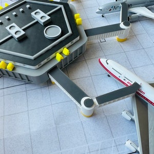May include: A model airport terminal with two airplanes docked. The terminal is gray with yellow accents and has a black roof. The airplanes are white with red and blue accents. One airplane has the text "American" on the side and the other has the text "Asiana" on the side.