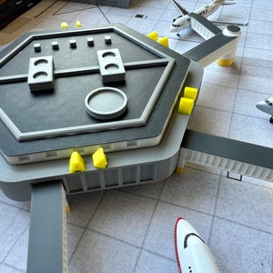 May include: A gray and white model airport terminal with three connecting jet bridges. The terminal has a hexagonal roof with a circular control panel and several rectangular buttons. There are three airplanes parked at the gates, two white and one red and white.