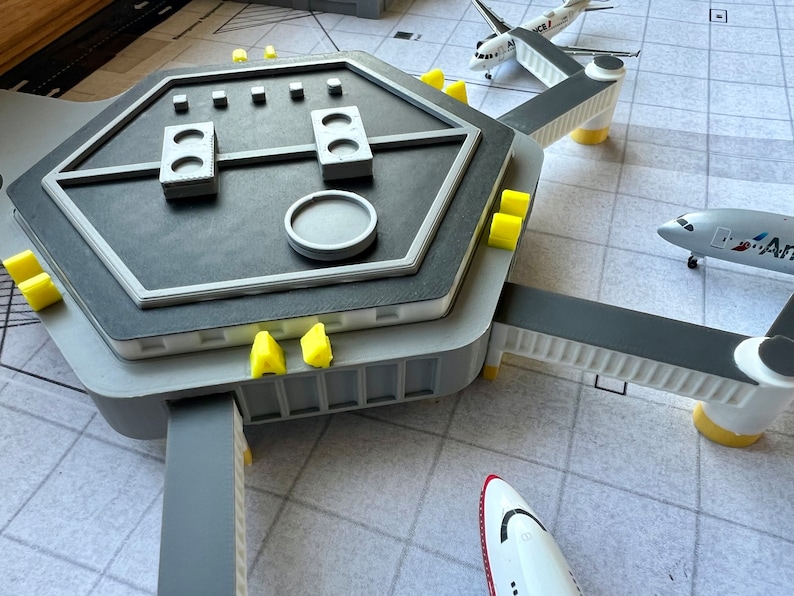May include: A gray and white model airport terminal with three connecting jet bridges. The terminal has a hexagonal roof with a circular control panel and several rectangular buttons. There are three airplanes parked at the gates, two white and one red and white.