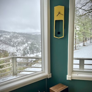May include: A yellow wooden pendulum clock with black hands hangs on a teal wall. The clock is in a room with two windows overlooking a snowy landscape. A record player and speaker are on a wooden table in the foreground.