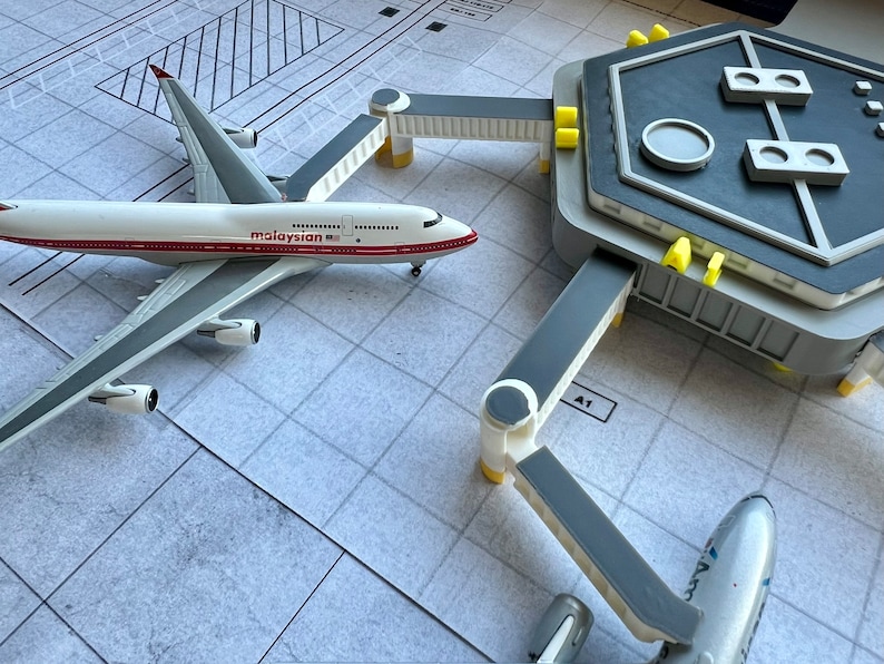 May include: A white airplane model with the word "Malaysian" printed on the side, parked at a gray airport terminal with a gray and yellow gate.