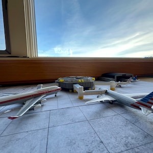 May include: A model airport terminal with three toy airplanes. A white and red airplane with "malaysian" written on the side, a white airplane with a blue and red American flag on the tail, and a smaller white airplane with a red stripe.