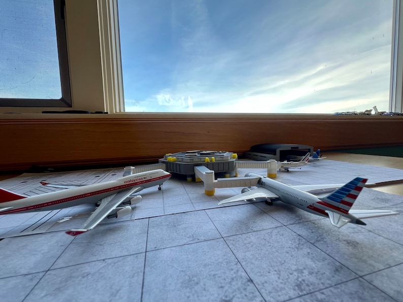 May include: A model airport terminal with three toy airplanes. A white and red airplane with "malaysian" written on the side, a white airplane with a blue and red American flag on the tail, and a smaller white airplane with a red stripe.