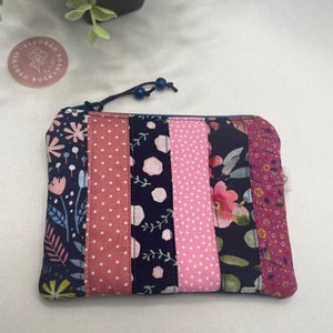 Pode incluir: Uma pequena bolsa retangular com zíper e design patchwork. A bolsa é feita de diferentes tecidos com padrões florais, bolinhas e listras em tons de rosa, azul e branco. O zíper é azul e tem um puxador de tassel azul. A bolsa tem uma etiqueta que diz "Algodão Rosa".
