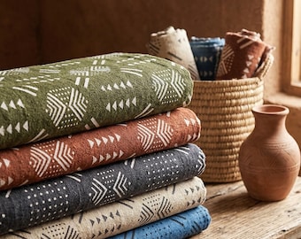 May include: A stack of folded textiles in shades of green, rust, gray, tan, and blue, each with a white geometric pattern. A woven basket holds rolled textiles, and a terracotta vase sits nearby on a rustic wooden surface.