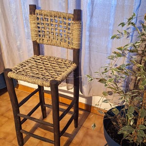 May include: A wooden chair with a woven seat and back. The chair is brown and has a rustic look. It is sitting in front of a window with sheer curtains. A potted plant is in the background.