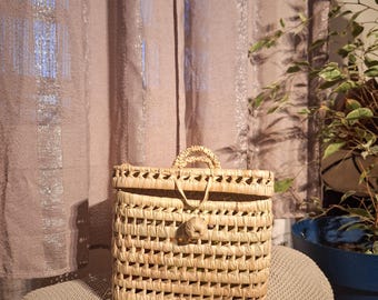 May include: A woven basket with a rectangular shape and a handle. The basket is made of light brown natural fibers and features a decorative closure. The background includes a curtain and a plant in a blue pot.