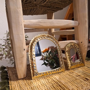 Puede incluir: Dos espejos arqueados dorados con bordes decorativos sobre una estera de paja tejida. El espejo más grande refleja un ramo de flores.