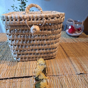 Puede incluir: Una cesta de mimbre tejida con tapa y asa. La cesta es de color beige natural y tiene un pomo redondo en la tapa. La cesta está sobre una estera de mimbre tejida.