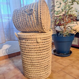 May include: A tall, cylindrical woven basket with a matching lid. The basket is made of natural-colored woven material and has a lid that sits on top. The basket has two handles and a latch to secure the lid. The basket is suitable for storage.