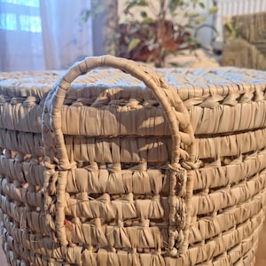 May include: Close-up of a woven basket with a rounded shape and a handle. The basket is made of natural-colored, dried plant fibers. The handle is arched and attached to the basket's rim. The weave is tight, creating a textured surface.