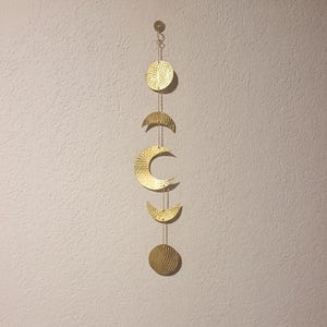Può includere: Un'applique da parete color oro raffigurante le fasi lunari. Il design include una luna piena, delle mezzelune e una falce di luna, tutti collegati da delicate catene. La superficie strutturata aggiunge interesse visivo a questo arredamento a tema celeste.