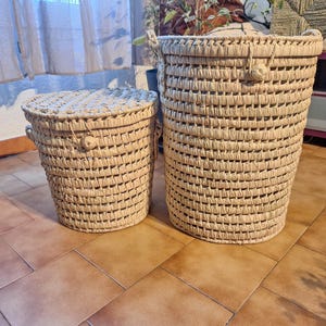 May include: Two woven baskets, one cylindrical and tall, the other shorter and round with a lid. Both are made of a light beige material and have a textured, woven pattern. The taller basket has handles. The baskets are suitable for storage.
