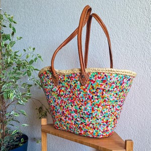Peut inclure: Un sac fourre-tout en crochet multicolore avec des anses en cuir marron. Le sac présente un motif moucheté de fils rouges, verts, bleus et jaunes. Le sac est posé sur une étagère en bois.
