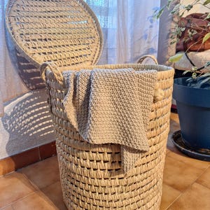 May include: A woven, cylindrical laundry hamper with a matching lid, partially open. The hamper is made of natural-colored woven material. A beige blanket is draped over the edge. The hamper is in a room with a tiled floor and a plant in the background.