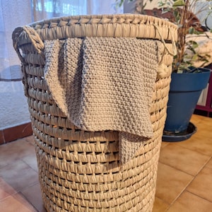 May include: A large, cylindrical woven basket made of natural fibers. A light gray knitted blanket is draped over the top edge. The basket has two handles and is sitting on a tiled floor.