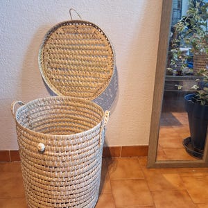 May include: A woven, cylindrical laundry basket with a matching lid. The basket is made of light-colored natural fibers, featuring two handles and a small knob on the side. The lid is round with a handle. The basket is on a tiled floor.