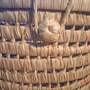 May include: Close-up of a woven basket made from natural fibers. The basket features a textured pattern with a handle and a decorative knot. The basket is a light brown color.