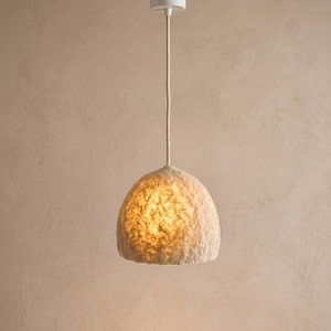 May include: A dome-shaped pendant light fixture with a textured, off-white fibrous shade. The lamp hangs from a single, straight, light-colored cord attached to a white ceiling mount. The interior emits a warm, soft, diffused golden glow.
