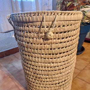 May include: A woven basket with a cylindrical shape, crafted from natural-colored straw. The basket features a textured, open weave design, with a sturdy handle and a decorative knot detail. The basket is suitable for storage or decorative purposes.