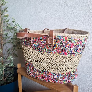 May include: A woven straw tote bag with a colourful sequin design around the top and bottom. The bag has brown leather handles and a neutral-coloured interior lining. The bag is sitting on a wooden shelf.