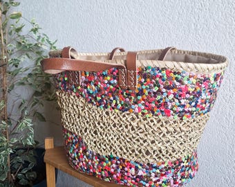May include: A woven straw tote bag with a colorful sequin design around the top and bottom. The bag has brown leather handles and a neutral-colored interior lining. The bag is sitting on a wooden shelf.