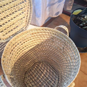 May include: A set of woven baskets in natural tones. The large, round basket has two handles and a textured weave. The lid of another basket is visible. The baskets are suitable for storage or decoration.