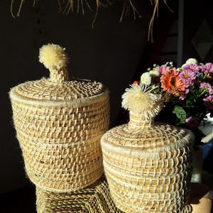 May include: Two woven baskets with lids. The baskets are cylindrical with a light brown color and a textured weave. Each lid has a fluffy, light yellow pom-pom on top. The baskets are sitting on a woven mat.