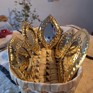 Puede incluir: Una cesta tejida con un borde dorado contiene un conjunto de seis pequeños espejos redondos de color dorado. Los espejos están dispuestos en círculo alrededor del centro de la cesta.