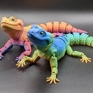 May include: Two articulated toy lizards, one orange and one blue with green, yellow, and purple accents. The lizards are posed on a black surface.