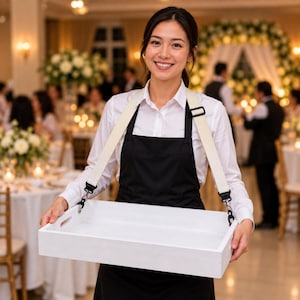 May include: A rectangular white wooden serving tray with integrated side handles, suspended by two thick beige canvas straps with black metal hardware clips, worn over the shoulders to allow for hands-free carrying of items in a professional setting.