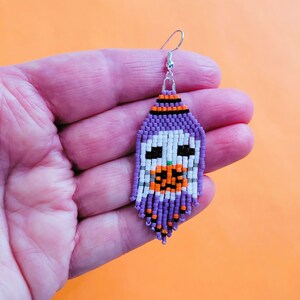 K&ouml;nnte beinhalten: Ein Perlenohrring mit einem Halloween-Geister-Design. Der Ohrring hat eine lila Oberseite, ein wei&szlig;es Gesicht und einen orangefarbenen Mund. Der Geist hat schwarze Augen und eine gr&uuml;ne Nase. Der Ohrring wird von einem silbernen Haken gehalten.