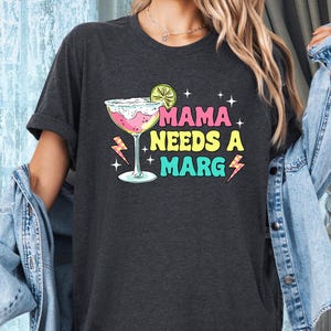 May include: A dark grey t-shirt with the text "Mama Needs a Marg" in colorful letters. A cartoon margarita glass with a lime wedge is next to the text.