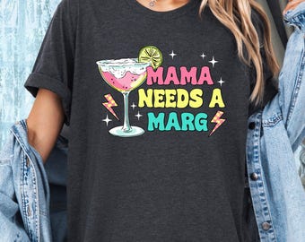 May include: A dark grey t-shirt with the text "Mama Needs a Marg" in colorful letters. A cartoon margarita glass with a lime wedge is next to the text.