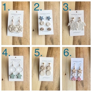 May include: Six pairs of handmade polymer clay earrings with various beach-themed designs, including seashells, starfish, pineapples, and turtles. The earrings are displayed on white cards with the brand name "HANNAH KATE" printed on them.