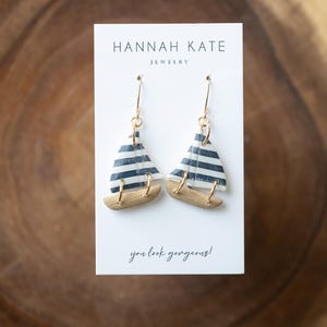 May include: A pair of sailboat earrings with gold-colored hooks. The sails are white with navy blue stripes, and the hulls are a light brown. The earrings are displayed on a white card with the text "HANNAH KATE JEWELRY" and "you look gorgeous!"