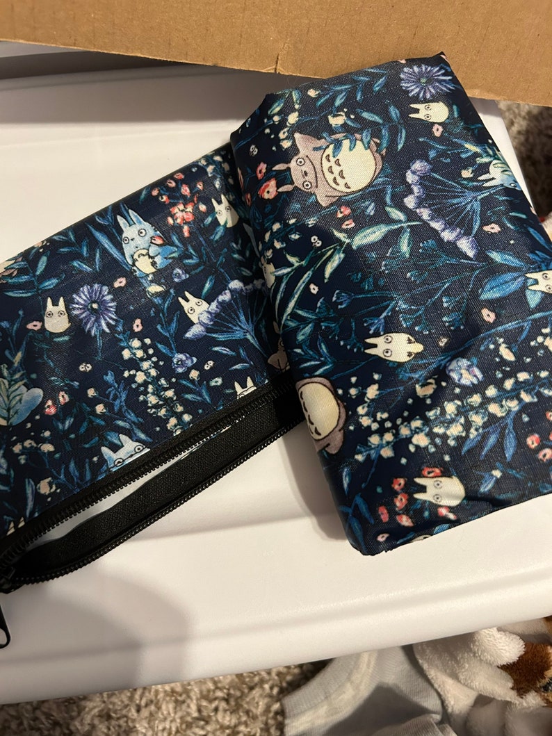May include: Two blue reusable shopping bags with a floral pattern and cartoon characters. The bags are folded and have a zipper closure.