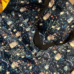 May include: A blue tote bag with a floral pattern featuring a cartoon character. The character is a grey and white creature with large ears and a fluffy tail. The bag has black straps.