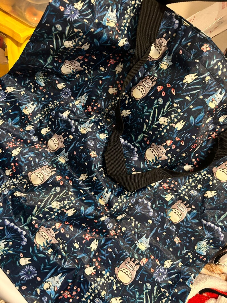 May include: A blue tote bag with a floral pattern featuring a cartoon character. The character is a grey and white creature with large ears and a fluffy tail. The bag has black straps.