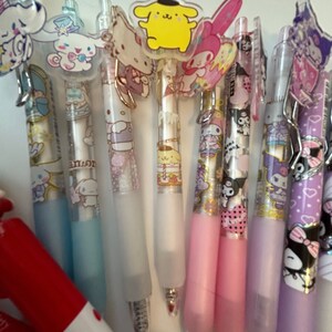 Op de afbeelding: Een verzameling van tien kleurrijke pennen met doorzichtige kunststof cilinders en verschillende cartoonfiguurtjes als toppers. De toppers omvatten Hello Kitty, My Melody, Little Twin Stars en een geel personage met een zwarte hoed. De pennen hebben een glinsterende, glimmende uitstraling.
