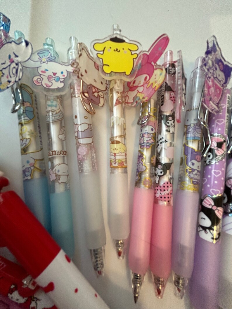 Op de afbeelding: Een verzameling van tien kleurrijke pennen met doorzichtige kunststof cilinders en verschillende cartoonfiguurtjes als toppers. De toppers omvatten Hello Kitty, My Melody, Little Twin Stars en een geel personage met een zwarte hoed. De pennen hebben een glinsterende, glimmende uitstraling.