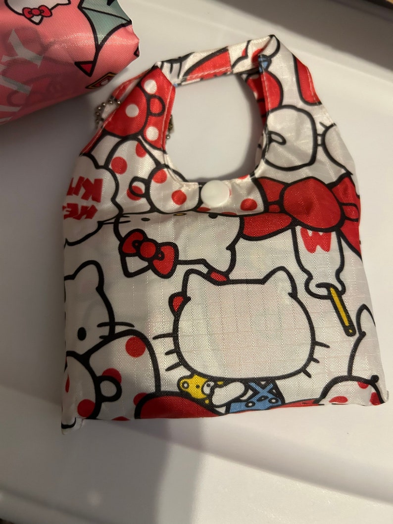 May include: A white reusable shopping bag with a red Hello Kitty design. The bag has red polka dots and a red Hello Kitty face with a red bow. The bag has a white handle and a white button.